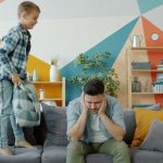 Stressed dad suffering from headache while naughty child beating him with pillow jumping on sofa having fun in apartment. Childhood and parenting concept.