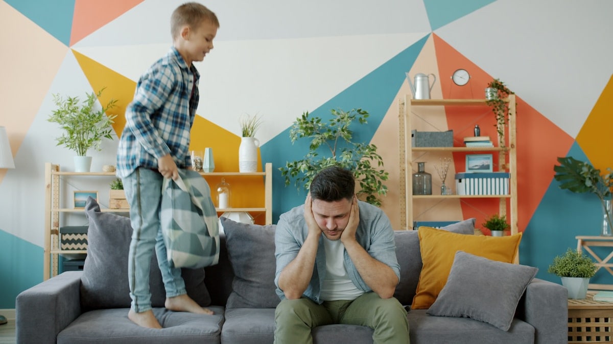 Stressed dad suffering from headache while naughty child beating him with pillow jumping on sofa having fun in apartment. Childhood and parenting concept.